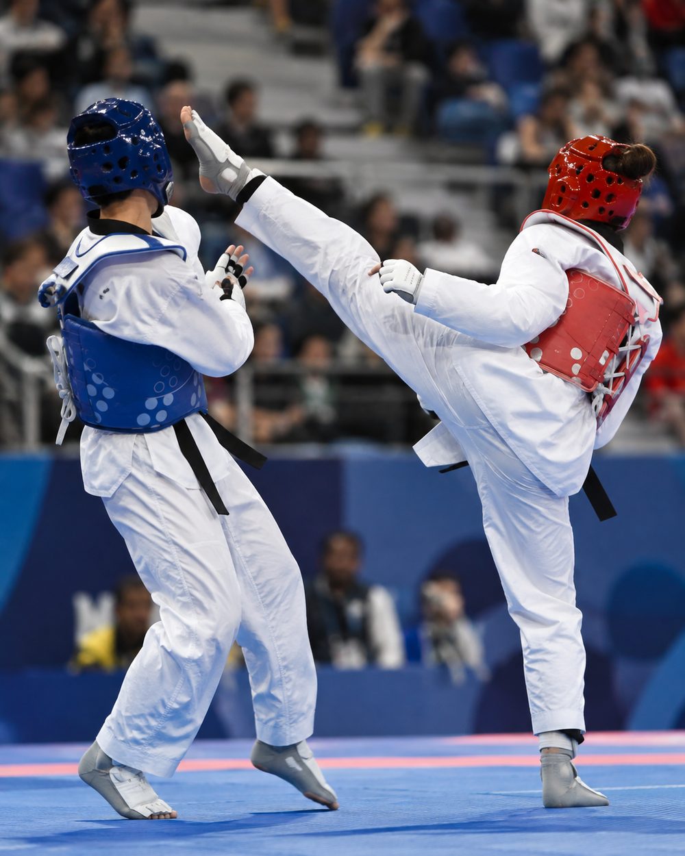 Müsabaka minderinde mavi ve kırmızı koruyucu ekipmanlı iki taekwondo sporcusu; kırmızı köşedeki sporcu yüksek tekme atıyor.